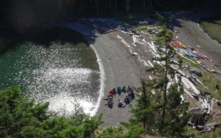 From an aerial point of view, a group of people sit in a circle on a beach. Kayaks and driftwood are also beached nearby.